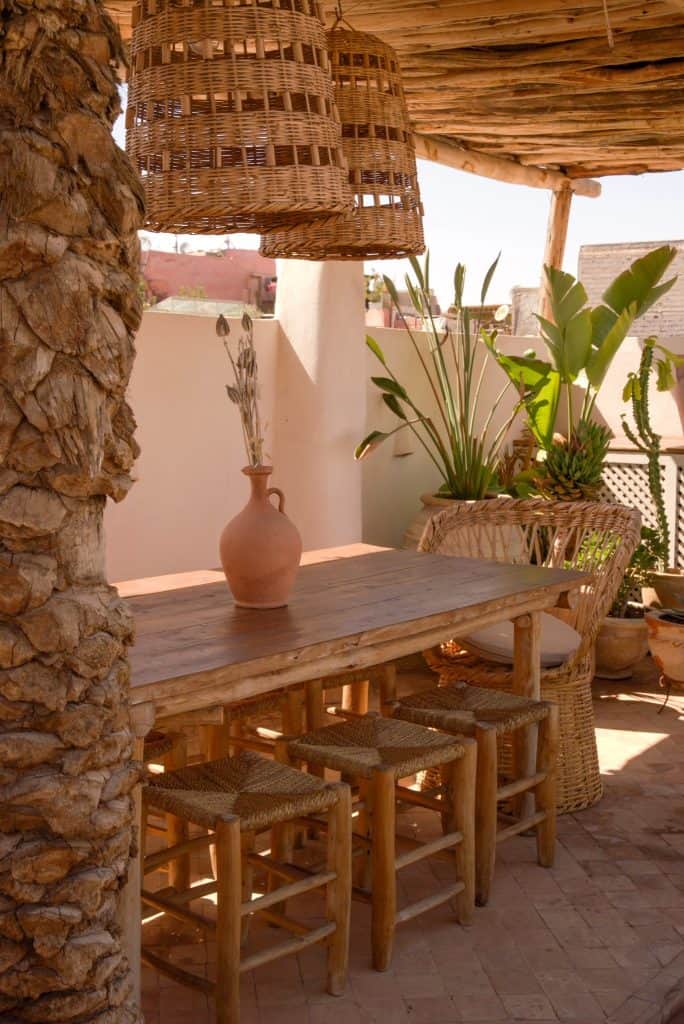 Marrakech rooftop dining nook with a rustic wooden table and woven rope stools, oversized rattan basket pendant lights, a terracotta jug holding dried stems, and tropical plants framing a stone column.
