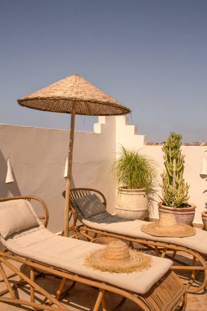 Marrakech riad rooftop with two rattan sun loungers topped with cream cushions and woven straw hats, shaded by a tall conical reed parasol, alongside potted cacti and grasses against a white tadelakt wall.