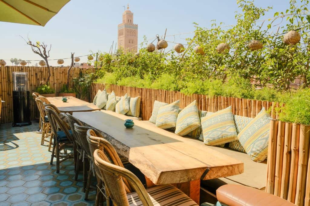 Marrakech rooftop terrace with a long live-edge wooden table and rattan chairs, banquettes piled with patterned blue and yellow cushions, bamboo fencing, and a view of the Koutoubia Mosque minaret rising above the leafy garden boundary.