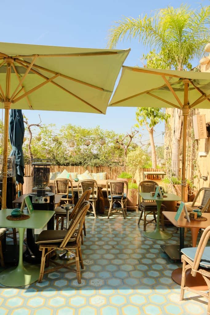Marrakech rooftop café with chartreuse umbrellas shading rattan chairs and small bistro tables, hexagonal blue patterned floor tiles, bamboo fencing, and tall palms rising against a clear sky.