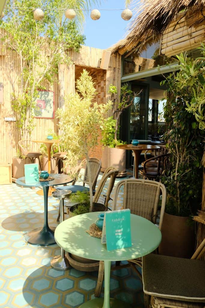 Marrakech rooftop café corner at Kabana with rattan chairs around small green and blue tables, turquoise hexagonal floor tiles, bamboo walls, potted olive trees, and woven ball lights strung overhead against a clear sky.