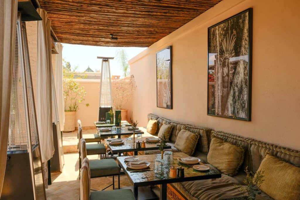 Marrakech rooftop restaurant with a slatted wooden ceiling, long banquettes piled with olive linen cushions, dark stone tables set for a meal, large framed photographic portraits on warm beige walls, and an open-air terrace beyond.