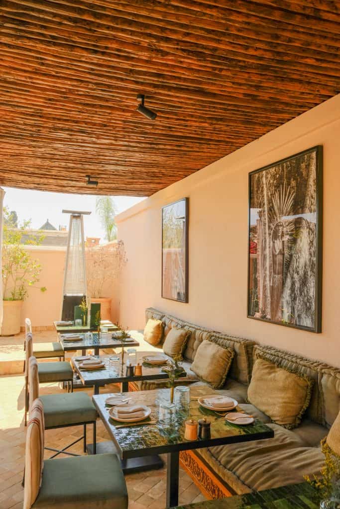Marrakech rooftop dining terrace with a slatted wooden ceiling, long banquettes piled with linen cushions, dark stone tables set for a meal, and large framed photographic prints on warm beige walls.