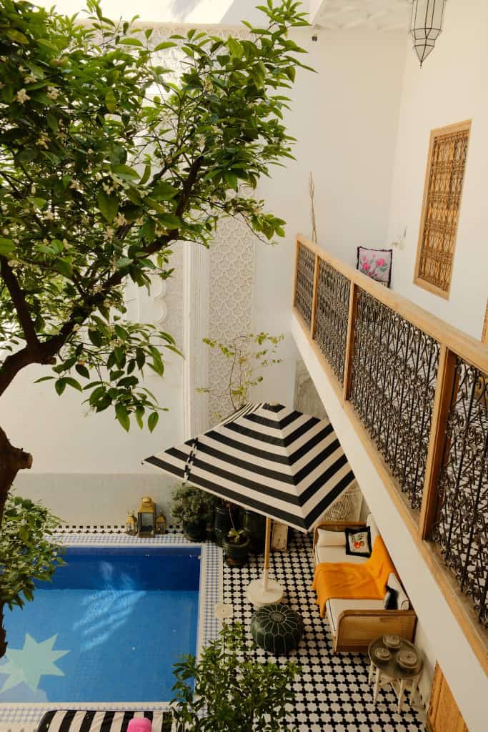 Image 1: "Overhead view of a Marrakech riad courtyard with a blue-tiled plunge pool, a black-and-white striped umbrella shading a rattan sofa with an orange throw, patterned floor tiles, and a flowering citrus tree reaching up past a wrought-iron balcony.