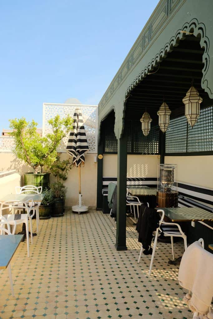 Marrakech riad rooftop terrace with green Moorish arches and brass lanterns, black-and-white striped umbrellas, white wrought-iron tables and chairs, patterned tile flooring, and small potted citrus trees along the edge.