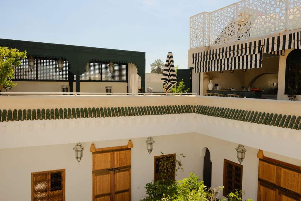 View across a Marrakech riad with white walls and green-tiled rooflines, looking up toward a rooftop terrace with black-and-white striped awnings, a black lattice pavilion, and palm trees against a clear sky.