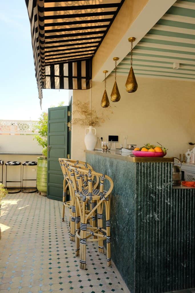 Marrakech riad rooftop bar with a textured green stone counter, rattan stools with black-striped trim, golden teardrop pendant lights, and a black-and-white striped awning above patterned tile floors.