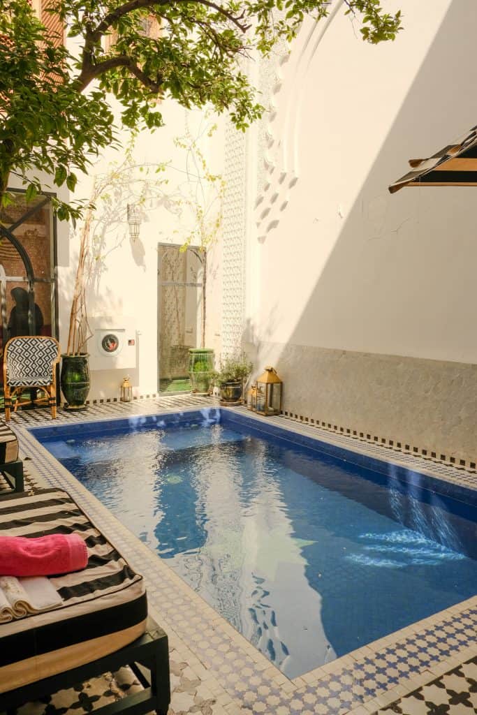 Marrakech riad courtyard with a small blue-tiled plunge pool, a striped sun lounger with red and white towels, brass lanterns and green ceramic pots along the edge, and dappled light filtering through an overhanging citrus tree.