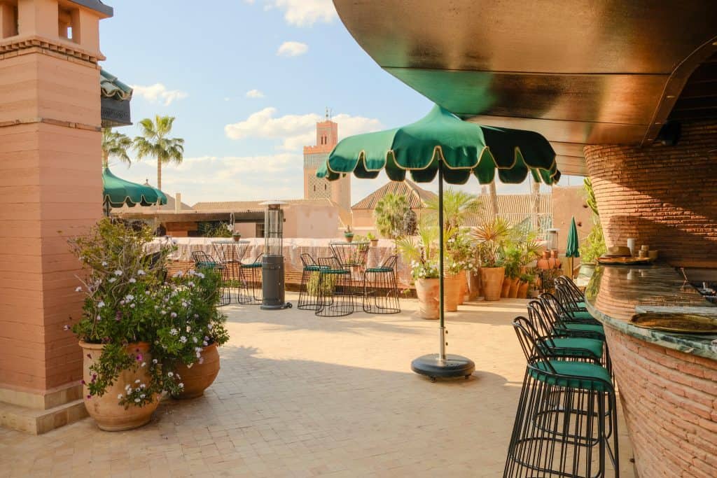 Marrakech rooftop bar with scalloped green umbrellas, black wire bar stools at a curved brick counter, terracotta planters of flowers, and a view across the medina to the minaret of the Koutoubia Mosque under a bright blue sky.