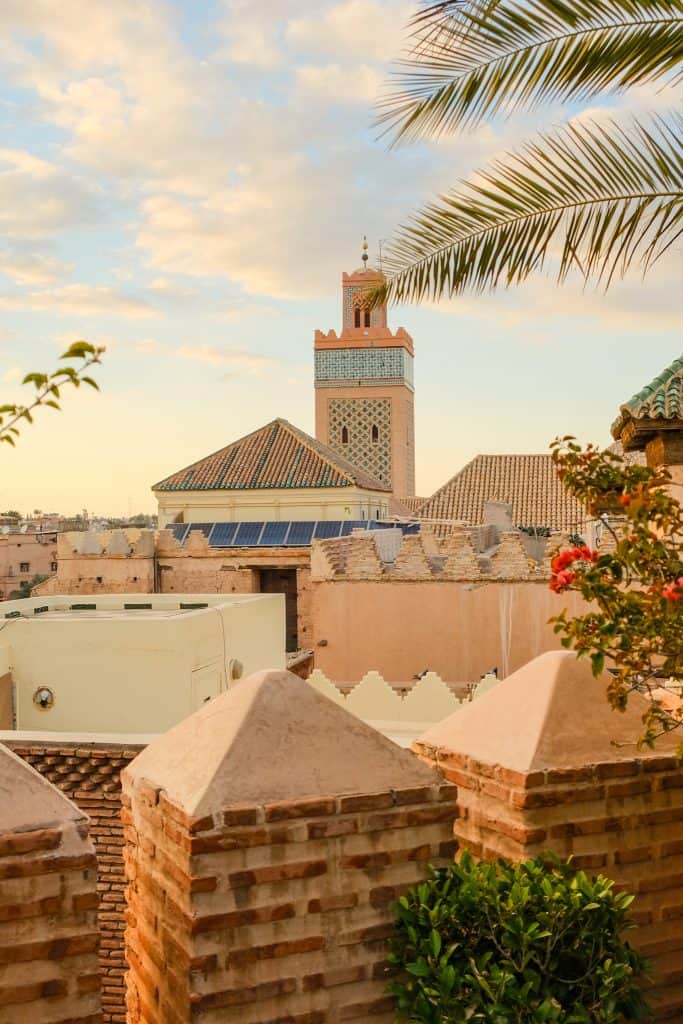 View from a Marrakech rooftop at golden hour, with terracotta brick crenellations in the foreground and the tiled minaret of a mosque rising above the medina rooftops under a pastel sky, framed by palm fronds and bougainvillea.