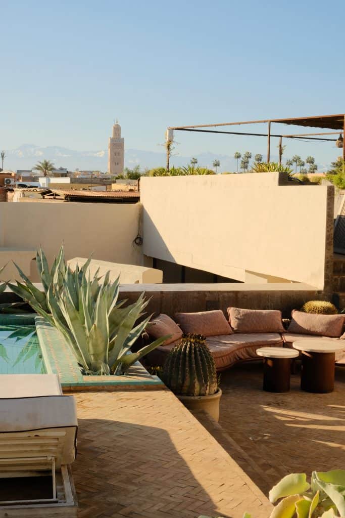 Marrakech riad rooftop terrace with a green-tiled plunge pool, a curved pink sofa scattered with cushions, agave and barrel cacti in the foreground, and a view across the medina to the Koutoubia Mosque minaret and the snow-capped Atlas Mountains.