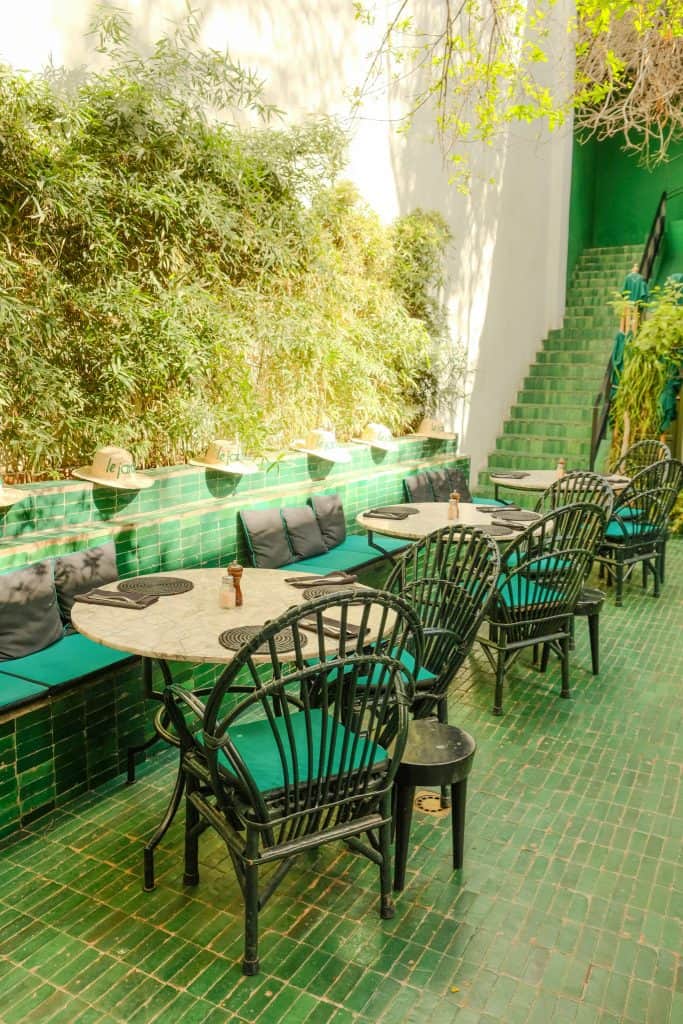 Marrakech garden restaurant with emerald green tiled banquettes and floors, dark green wicker chairs around marble bistro tables, straw sun hats lined up along the seating, and a white staircase leading up through bamboo and tropical greenery.