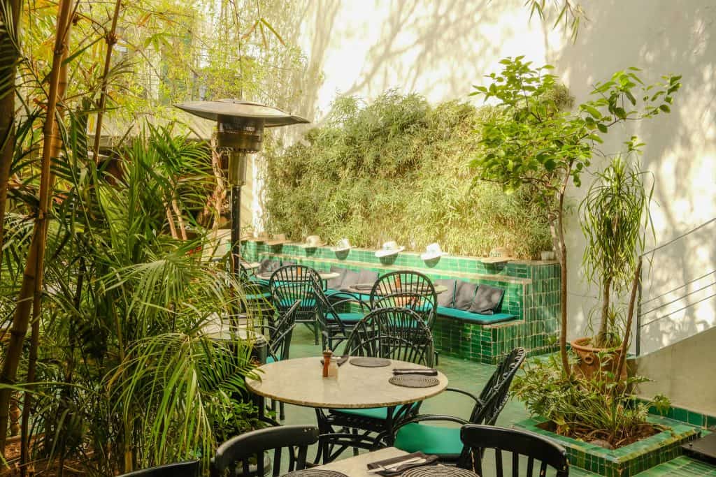 Lush Marrakech restaurant garden with emerald green tiled banquettes and floors, dark green wicker chairs around marble bistro tables, and tall bamboo and palms enclosing the space in dappled sunlight.