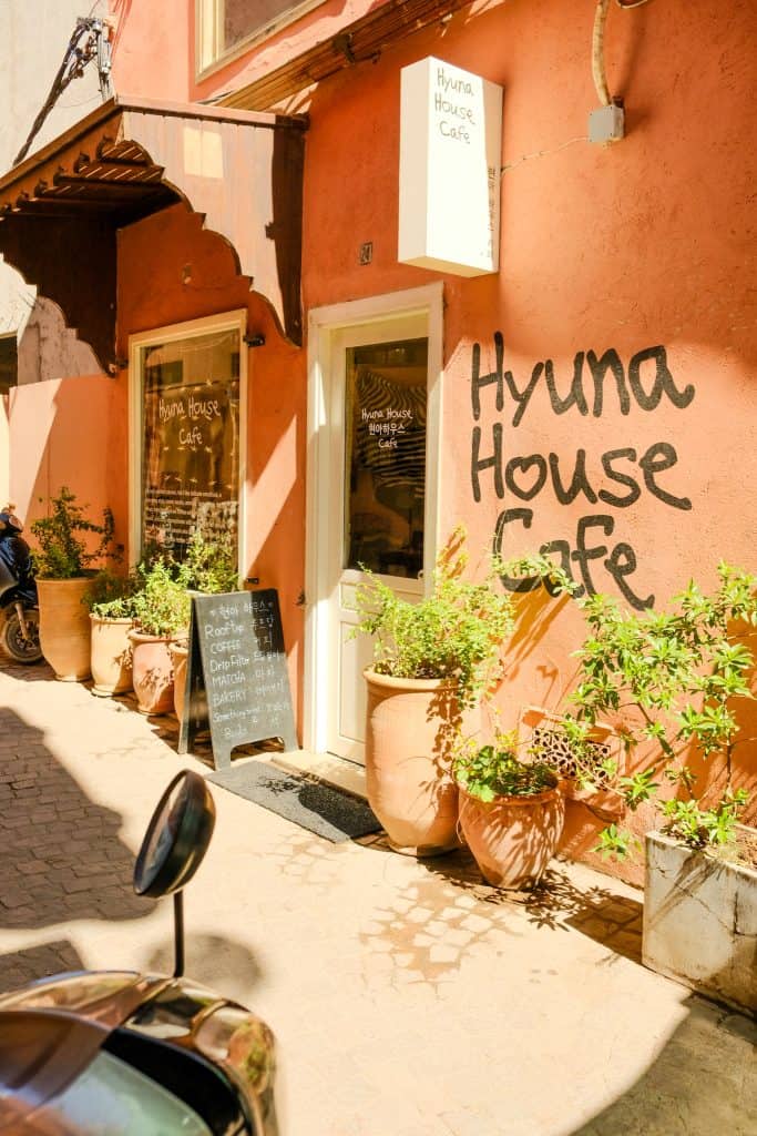 Exterior of Hyuna House Café in Marrakech, with its name hand-painted in large black script on a salmon-pink wall, a chalkboard menu and terracotta planters lining the entrance along a sunlit cobblestone alley.