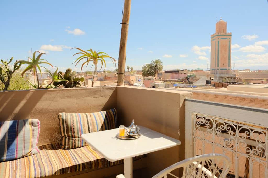 Marrakech rooftop café corner with a striped kilim-cushioned banquette, a small marble-topped table set with a silver teapot and glass of mint tea, and a panoramic view across the medina rooftops toward a tall sandstone minaret.