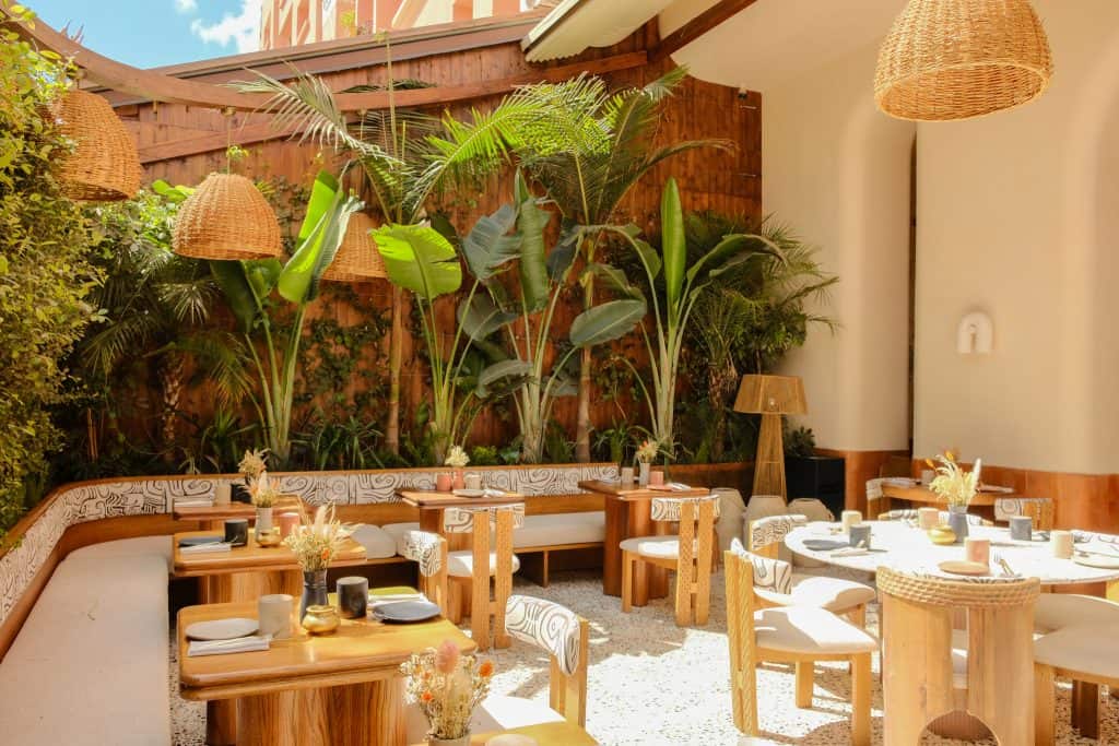 Marrakech open-air restaurant with light wood tables and curved chairs, banquettes upholstered in a black-and-white abstract pattern, oversized woven rattan pendants overhead, and a backdrop of tall banana plants and palms against a wooden wall.