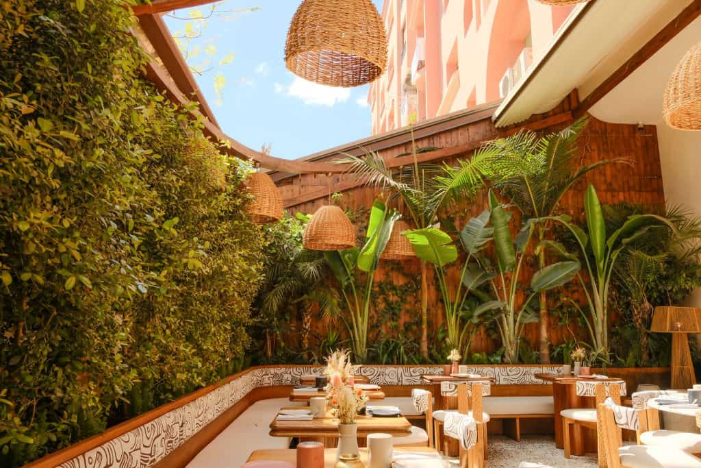 Marrakech open-air restaurant with banquettes upholstered in a black-and-white abstract pattern, warm wood tables and curved-back chairs, oversized woven rattan pendant lights overhead, and walls of lush greenery and tall banana plants enclosing the space under a bright blue sky.