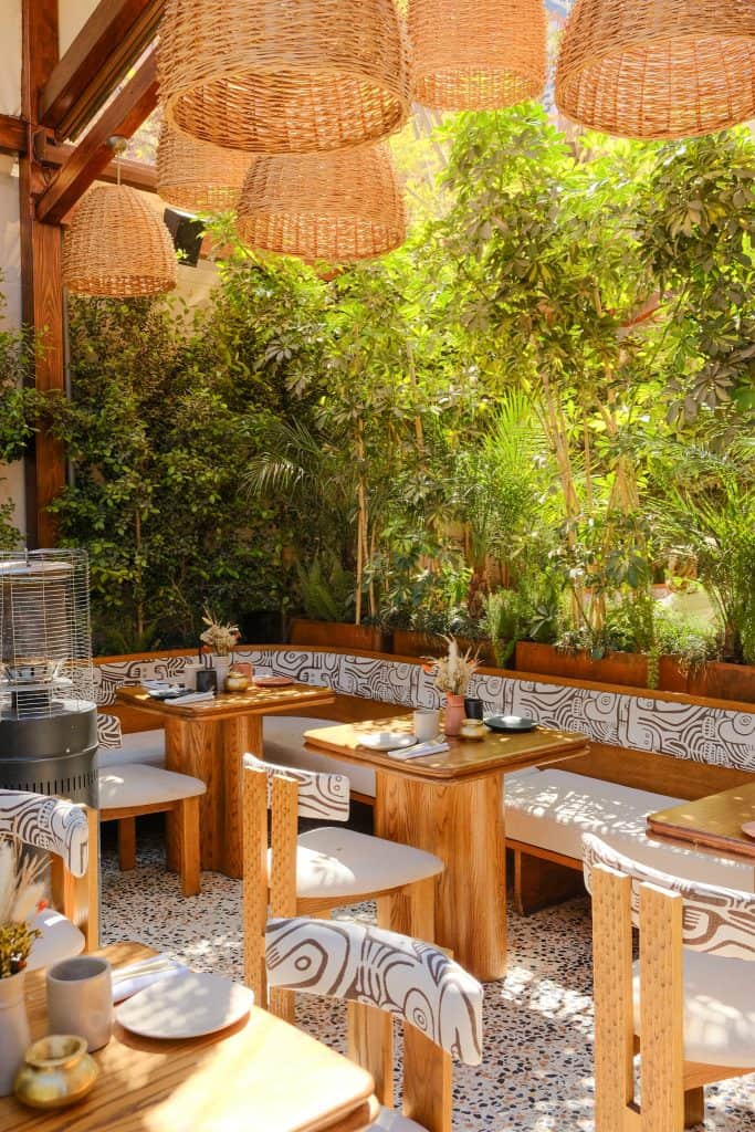 Marrakech garden restaurant with patterned black-and-white banquettes, light wood tables and chairs, oversized woven rattan pendant lights, and a backdrop of lush tropical foliage and bamboo.