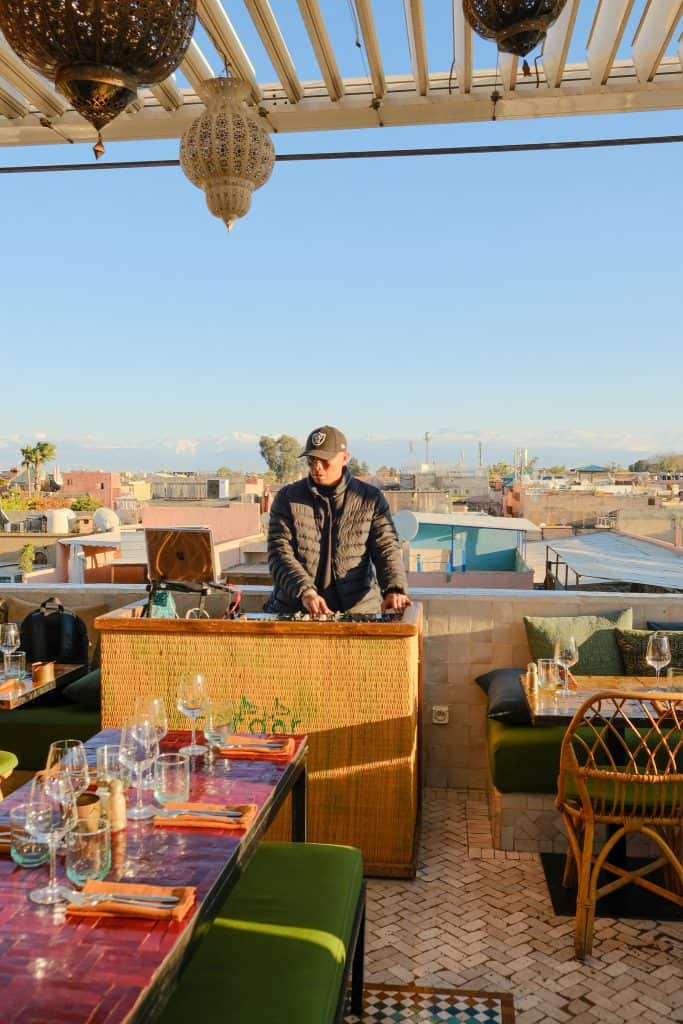 A DJ in a baseball cap and gilet plays a set at a woven bamboo booth on a Marrakech rooftop, with ornate hanging lanterns above, green banquettes and red-tiled tables in the foreground, and the snow-capped Atlas Mountains visible in the distance.