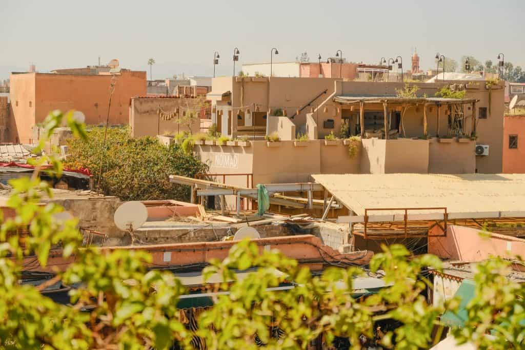 View across the Marrakech medina rooftops toward the Nomad restaurant terrace, with its pergola-shaded dining area perched above terracotta-toned buildings, satellite dishes, and a distant minaret on the horizon.