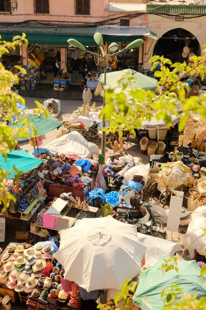 Aerial view of a busy Marrakech market square seen through tree branches, with stalls of colorful textiles, woven baskets, and straw hats sheltered under white and green parasols.