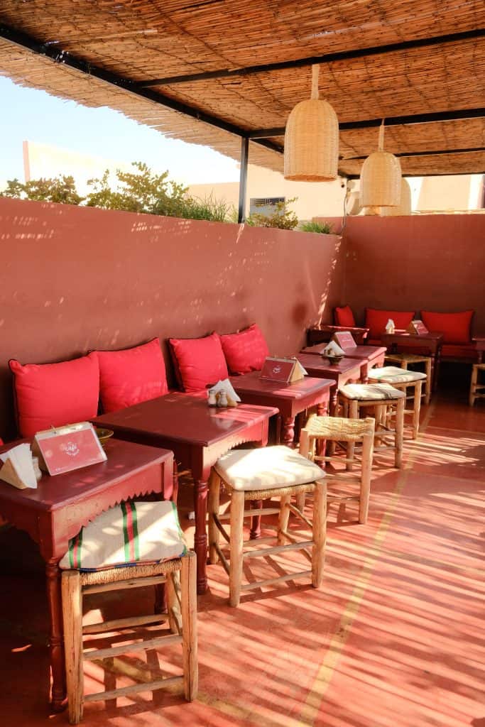 Quiet corner of a Marrakech rooftop café with terracotta floors and walls, dark wooden tables and stools topped with red cushions, woven rattan pendant lights, and a reed-slatted pergola overhead.