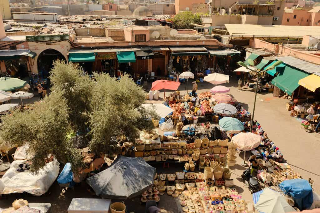 Elevated view of a bustling Marrakech market square filled with stalls of woven baskets, straw bags, and souvenirs sheltered under colorful parasols, with shopfronts and pink medina rooftops stretching into the distance.