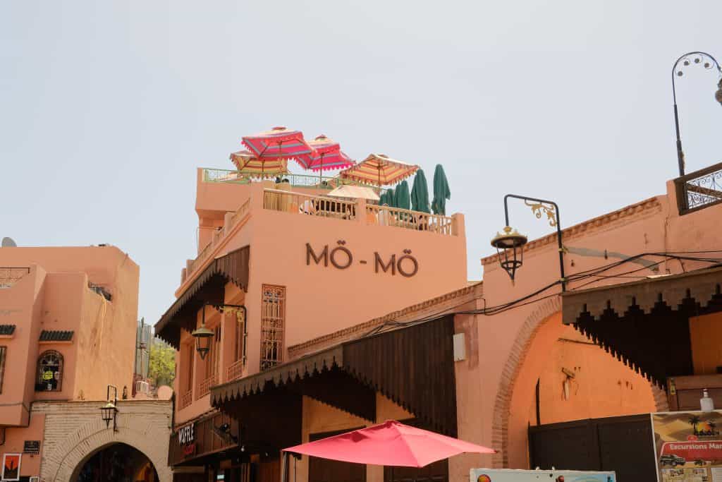 Exterior view of Mō-Mō restaurant in Marrakech, with colorful striped parasols visible on the rooftop terrace above a salmon-pink building, framed by the carved wooden eaves and brick archways of the surrounding medina.