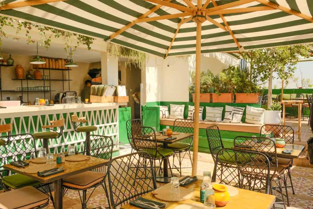 Marrakech rooftop café with a green-and-white striped umbrella, black wire chairs around small wooden tables, patterned encaustic floor tiles, a green-and-white latticed bar counter, and built-in benches with patterned cushions along a planted edge.