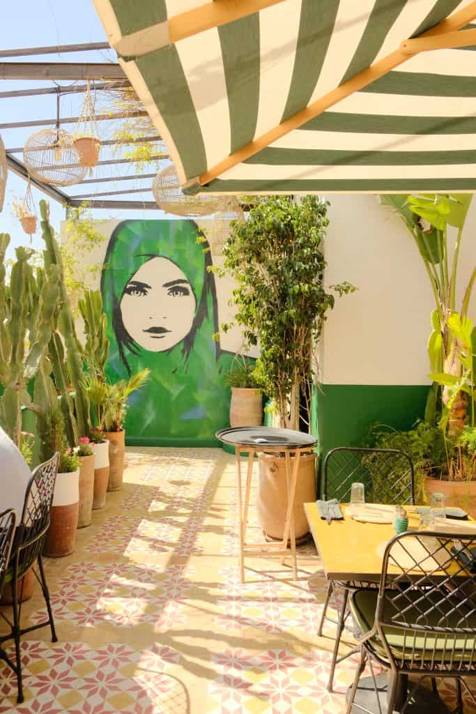 Marrakech rooftop café with patterned encaustic floor tiles, a large mural of a woman in a green headscarf on a white wall, hanging rattan lanterns under a green-and-white striped awning, and a black metal bistro table flanked by cacti and potted plants.