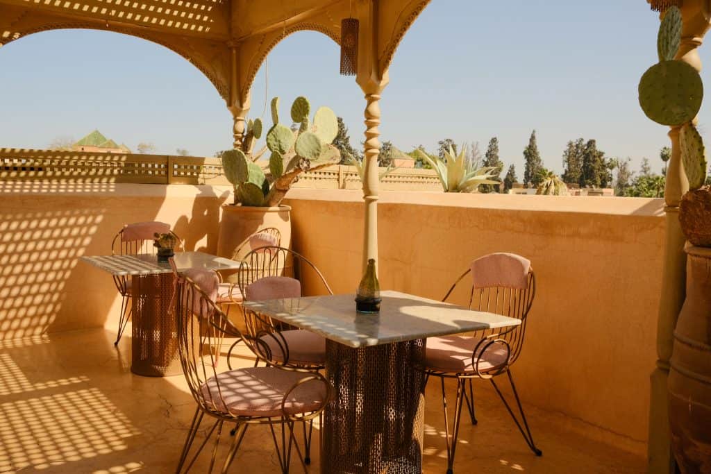 Marrakech rooftop terrace with carved peach-colored arches, a marble bistro table flanked by pink-cushioned wire chairs, prickly pear cacti in terracotta pots, and a sun-drenched view across rooftops to distant trees.