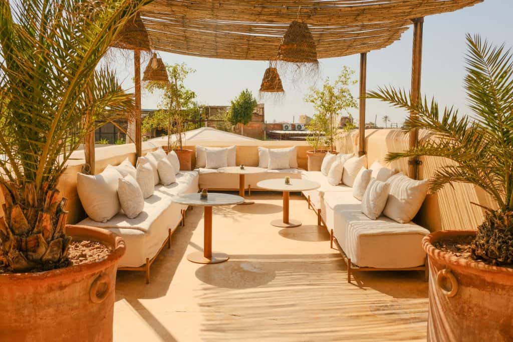 Marrakech riad rooftop lounge with long cream cushioned benches piled with linen pillows, low wooden tables, woven rattan pendant lights hanging from a reed-and-bamboo canopy, and terracotta planters of palms framing the space.