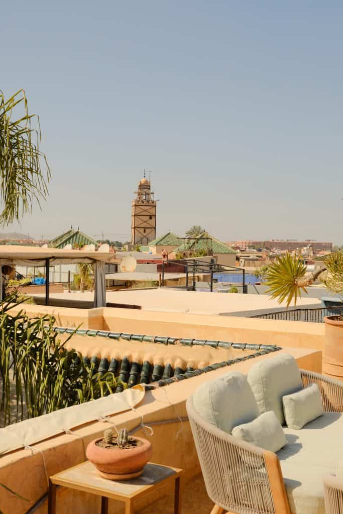 Marrakech rooftop seating area at golden hour, with a cream rattan sofa and pale blue cushions on a low wooden table, a green-tiled fountain edge, and a view across pink rooftops toward a tall stone minaret.