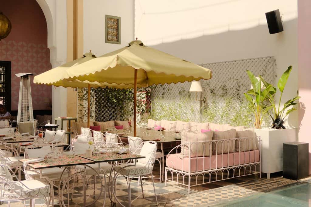Elegant Marrakech restaurant courtyard with scalloped olive green parasols, white wrought-iron chairs and daybeds with pink and striped cushions, mosaic-topped tables, and lush greenery climbing decorative metal screens beside a small pool with black-and-white checkered tiles.