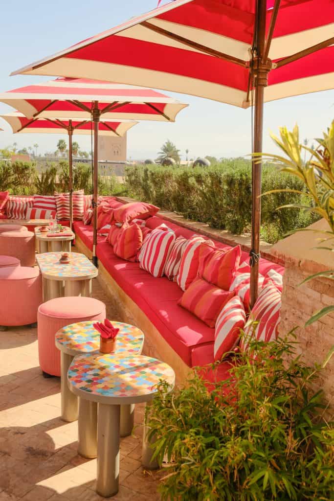 Marrakech rooftop terrace with red-and-white striped umbrellas shading long banquettes piled with pink and striped cushions, colorful mosaic-tiled side tables, and a view across rosemary hedges to the Koutoubia Mosque minaret in the distance.