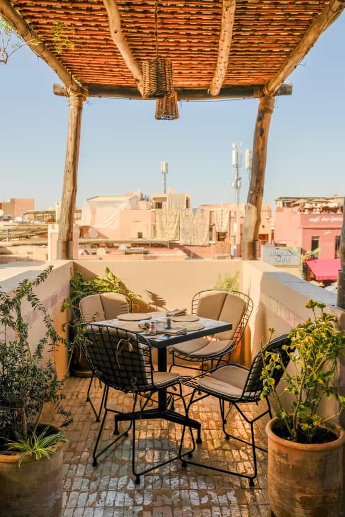 Intimate Marrakech rooftop corner with a small black wire table and chairs under a rustic reed-and-wood pergola, terracotta planters of greenery, and a view across the medina rooftops toward Café des Épices.
