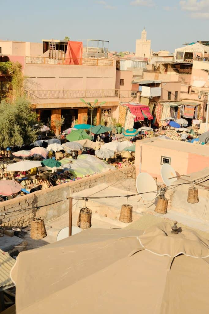 Elevated view across a Marrakech market square, with a sea of multicolored parasols and stalls below, surrounded by pink medina buildings and a slender minaret rising in the distance.