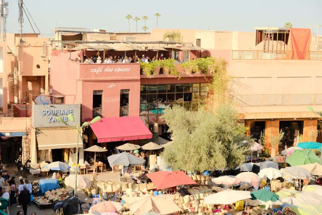 View across a Marrakech market square toward the pink façade of Café des Épices, with its rooftop terrace busy with diners overlooking the bustling stalls of woven baskets, parasols, and shoppers below.
