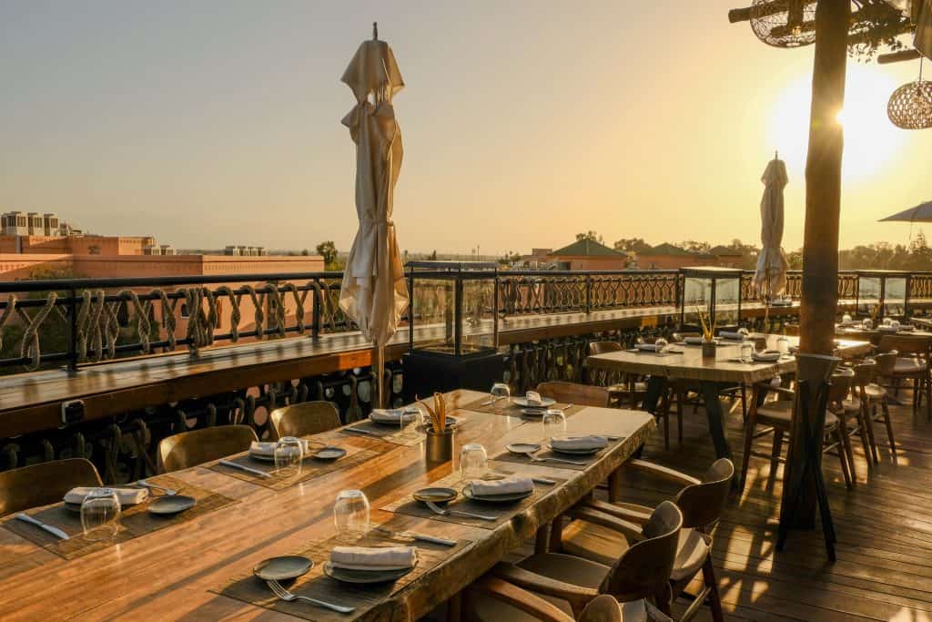 Marrakech rooftop restaurant at sunset, with long wooden tables set for dinner, closed cream parasols, and woven rope railings along the terrace edge, overlooking the city's pink rooftops bathed in golden light.