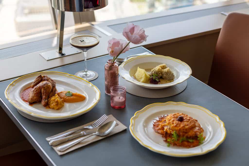 Table set with three savoury dishes at the Berlin TV Tower restaurant, including roasted meat, a creamy bowl dish, and a pastry-topped plate, alongside a coffee cocktail.