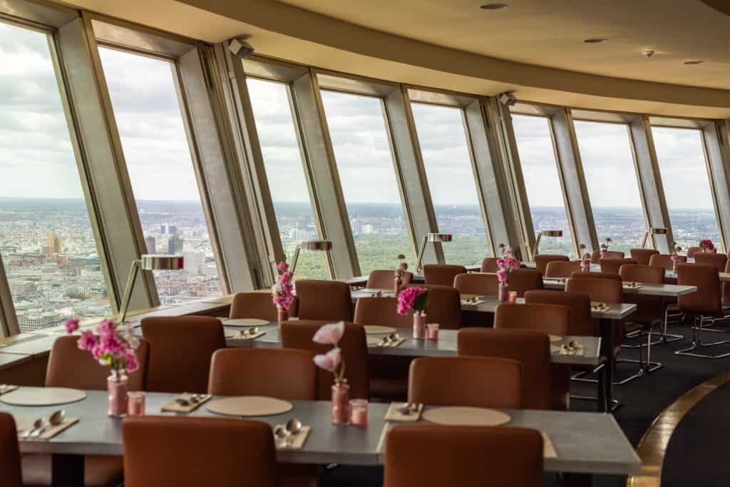 Curved dining room in the Berlin TV Tower's revolving restaurant, with rows of set tables, pink flower vases, and floor-to-ceiling windows overlooking the city.