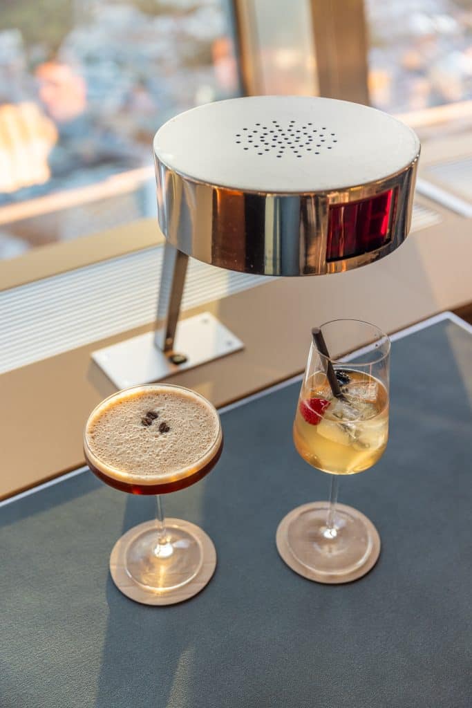 Two cocktails on a table at the Berlin TV Tower, an espresso martini and a garnished sparkling cocktail, with city light reflecting off the metal lamp above.
