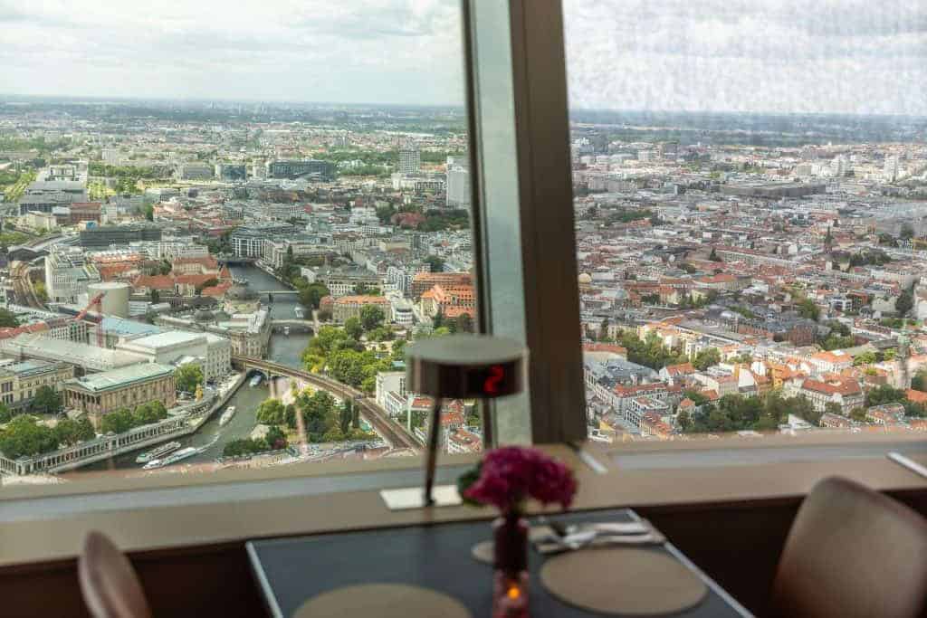 Panoramic view of Berlin's cityscape from a restaurant table at the TV Tower, with the Spree River, museum buildings, and red-roofed rooftops stretching to the horizon.