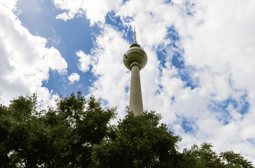 Berlin TV Tower rising above a line of trees against a partly cloudy sky.