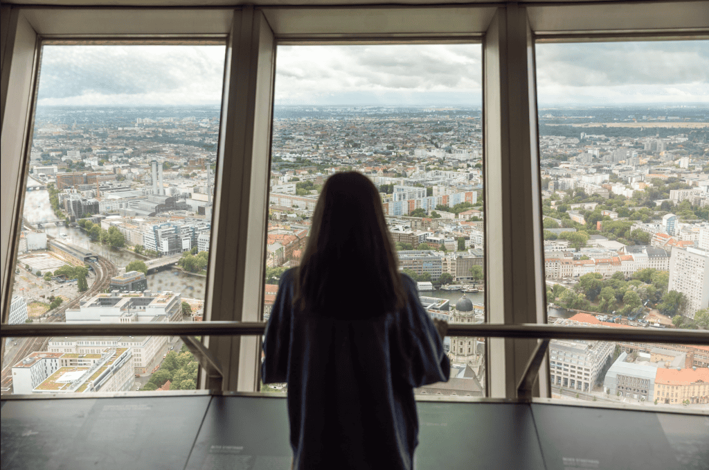 Woman viewed from behind looking out over Berlin's rooftops through the observation deck windows of the TV Tower.