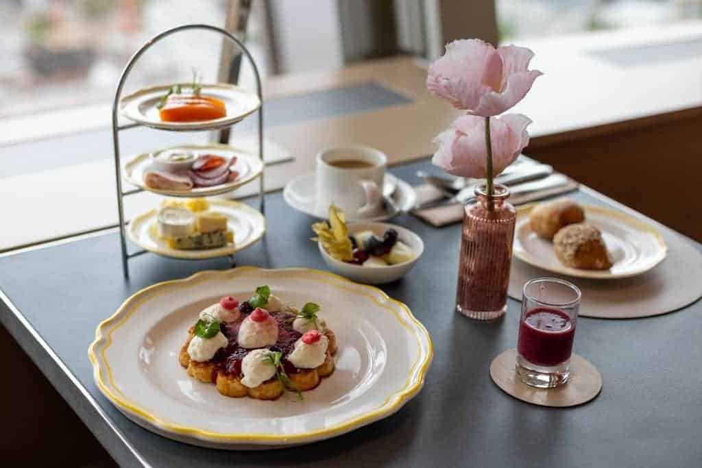Breakfast spread at the Berlin TV Tower restaurant with a waffle topped with berries and cream, a three-tier stand of savoury bites, coffee, pastries, and a berry juice shot.