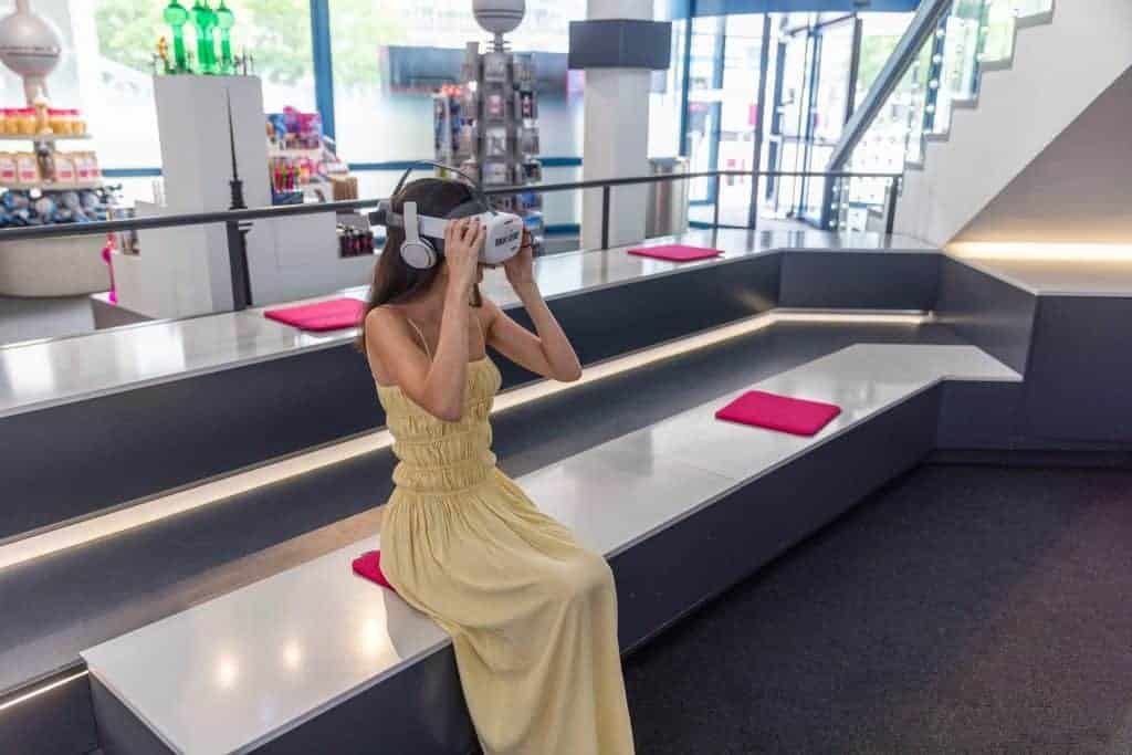 Woman in a yellow maxi dress wearing a VR headset, seated on a bench inside the Berlin TV Tower's visitor area with gift shop displays in the background.
